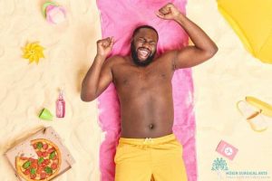 A man laying on the beach with pizza and other items.